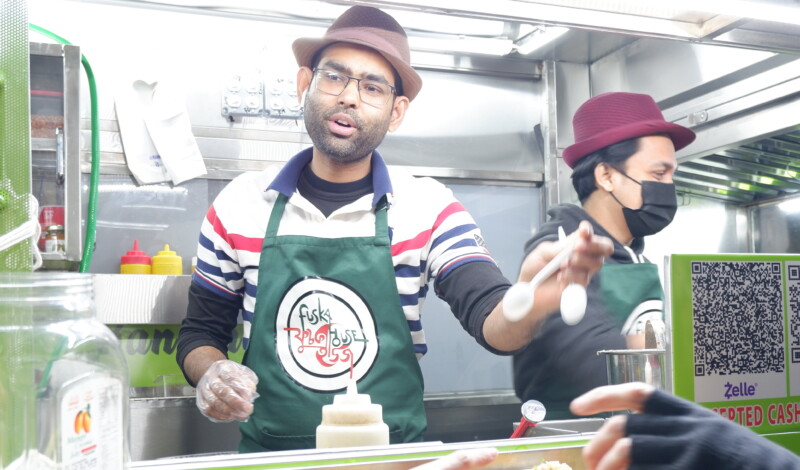 Vendor from Fushka House cart wearing a green apron and brown hat passes out bowls of food.